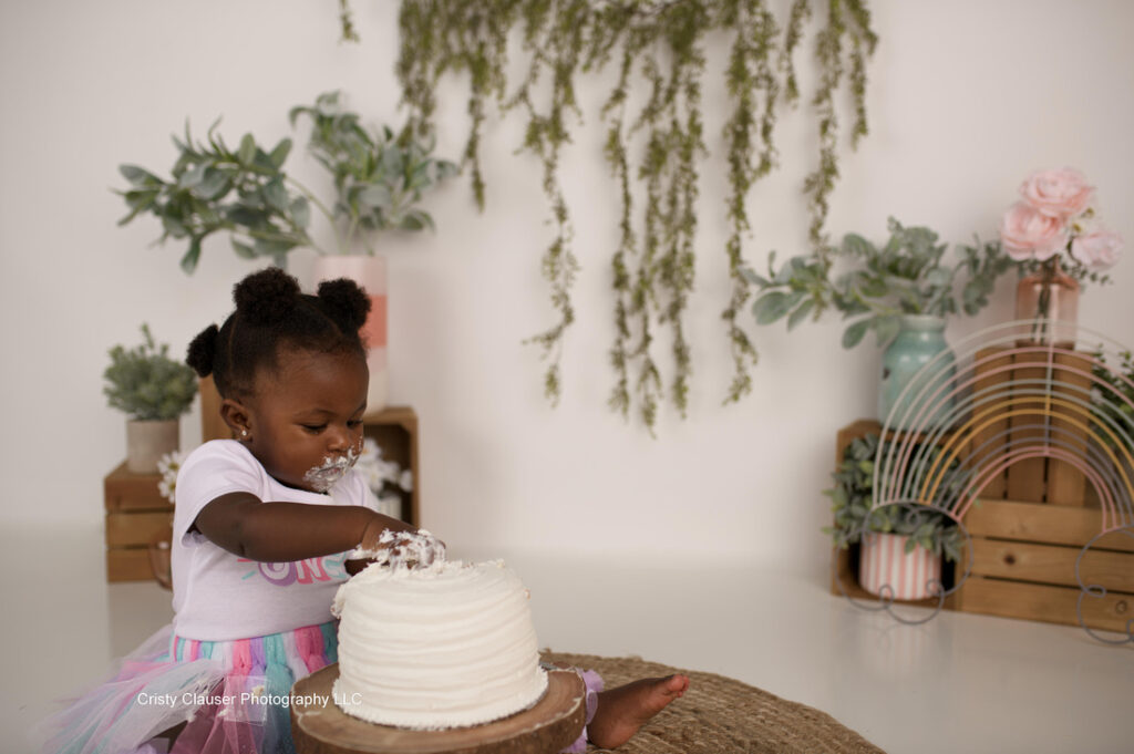 A toddler wearing a colorful tutu smashes a white cake with their hands. The background features potted plants, a hanging greenery decoration, and a decorative rainbow. The setting is bright and cheerful.