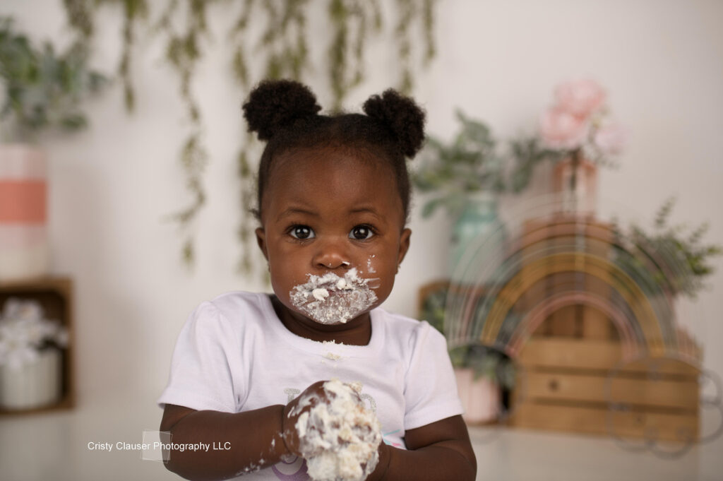 A toddler with two small buns in her hair holds a cupcake, with frosting on her face and hands. She is wearing a white shirt. The background features pastel decorations and hanging greenery.