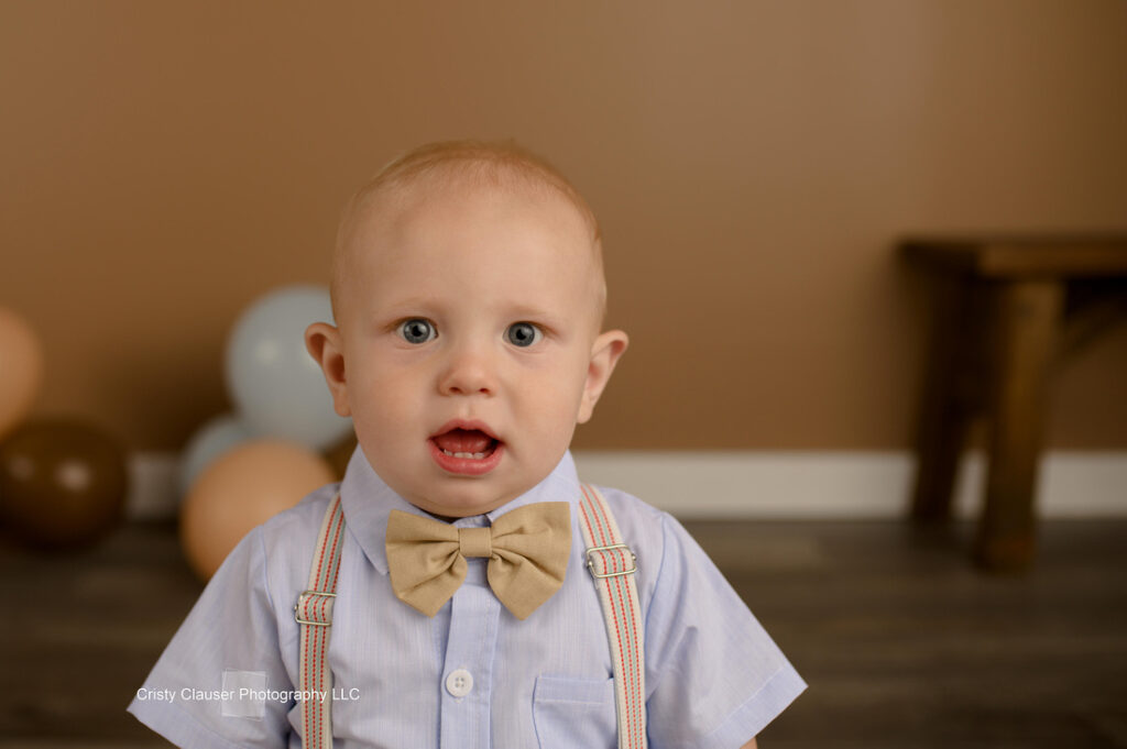 A toddler with short blonde hair is wearing a light blue shirt, tan bow tie, and suspenders. He stands indoors against a brown wall, with blurred balloons and a wooden stool in the background.
