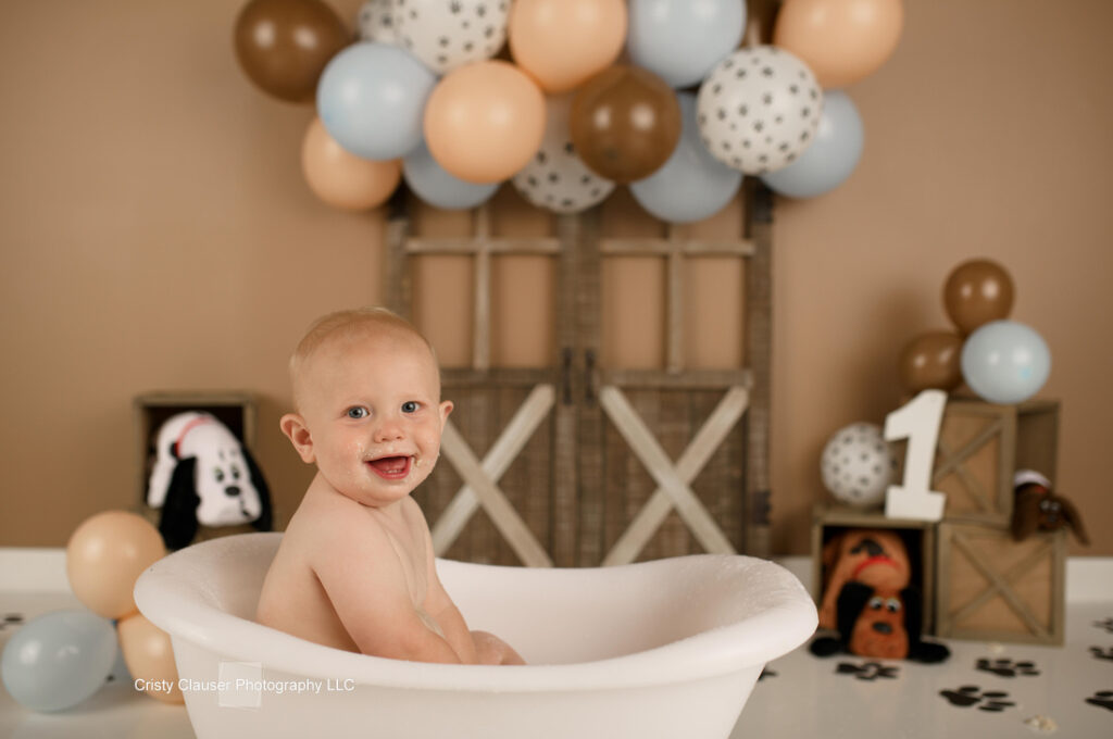A smiling baby sits in a white bathtub in front of a rustic backdrop with tan and blue balloons. To the side, there's a wooden crate with a large number 1 and a plush dog toy. The room has a playful, birthday-themed ambiance.