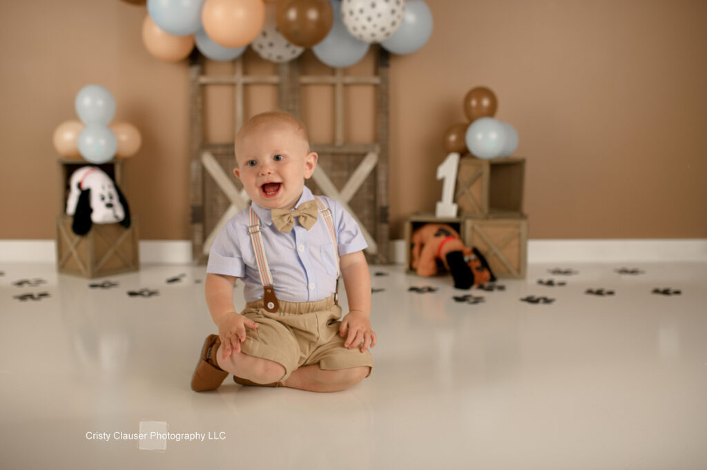 A baby wearing a bow tie and suspenders sits on the floor smiling. Behind him are balloons in blue, brown, and white. There are cubes, one with the number "1" on it, and a plush dog on the floor.