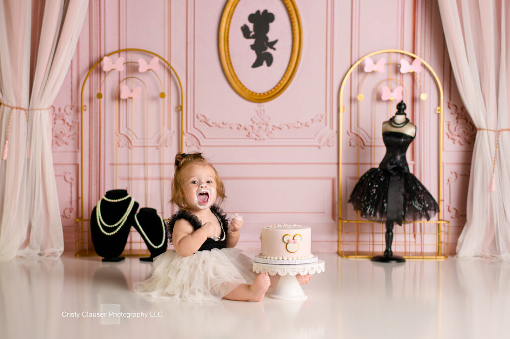 A toddler in a black and white tutu sits on the floor, smiling with cake frosting on her face. Behind her is a Minnie Mouse-themed cake on a stand. The room has pink walls with elegant decorations and mannequin displays.
