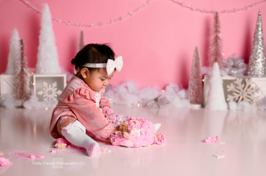 A baby wearing a pink outfit and white bow sits on the floor, playing with a pink cake. The background features pink, white, and silver holiday decorations, including trees and snowflakes.