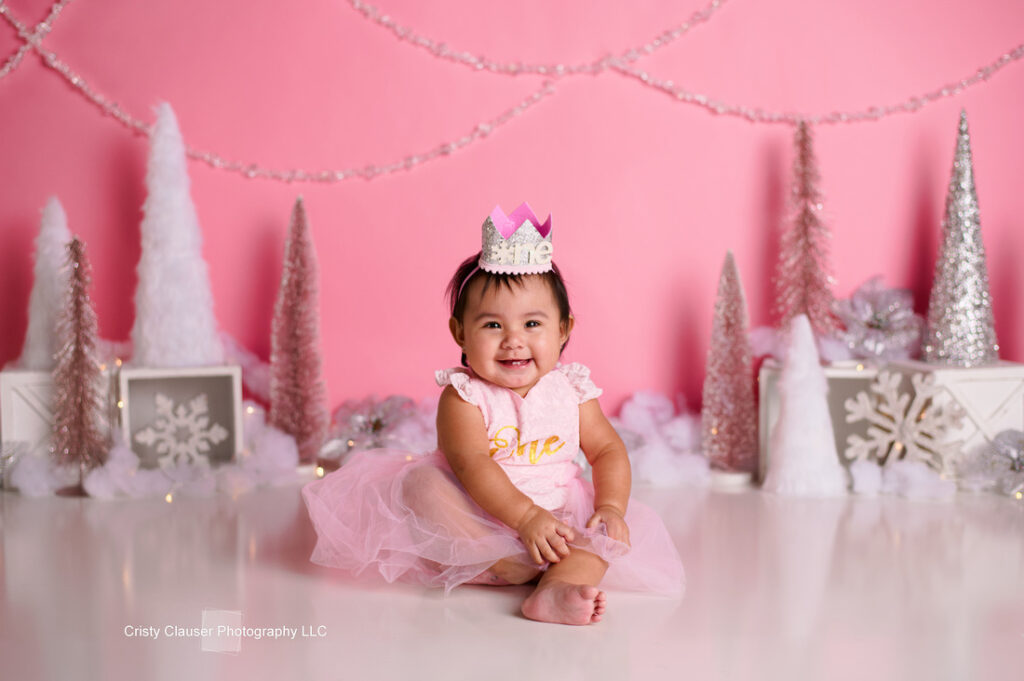 A baby girl wearing a pink dress and a small crown sits on the floor, smiling. She is surrounded by pink and white holiday decorations, including trees and snowflakes, against a pink background.