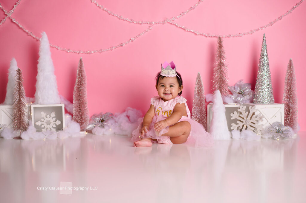A smiling baby wearing a pink dress and crown sits on a shiny floor. The background is decorated with pink and white Christmas trees, snowflakes, and festive garlands against a pink backdrop.
