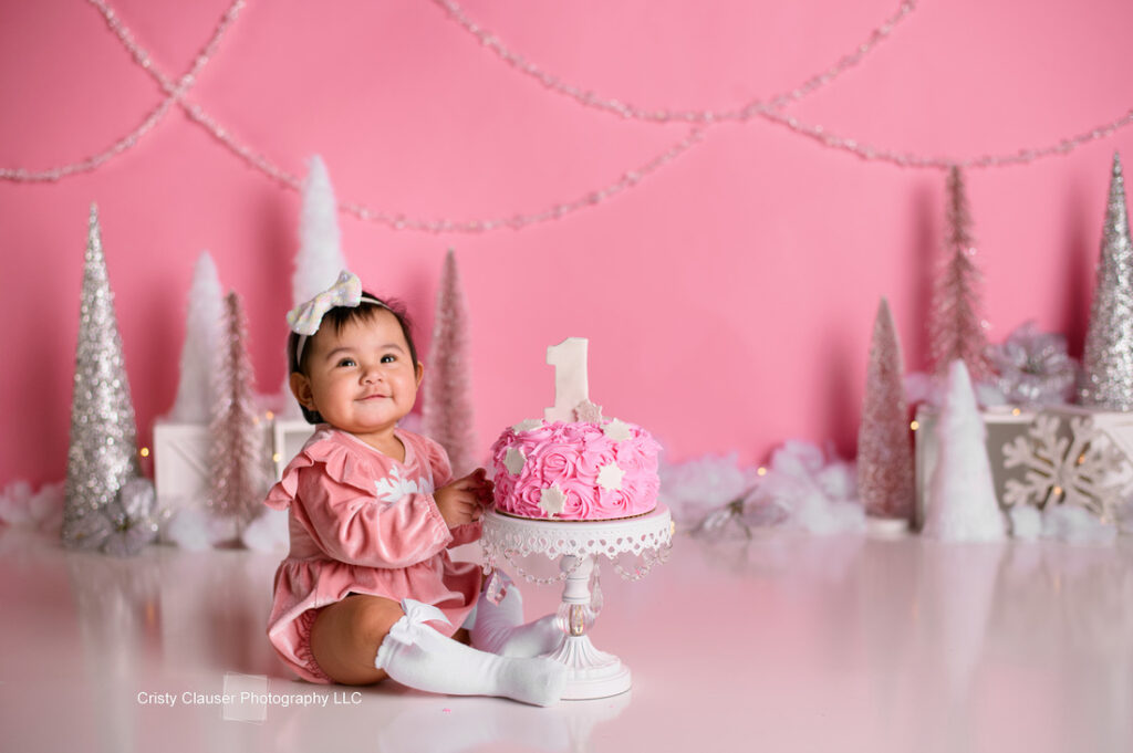 A baby wearing a pink dress and white knee-high socks sits next to a pink frosted cake with a "1" on top. The background features a pink wall adorned with silver and white decorative trees and garlands.