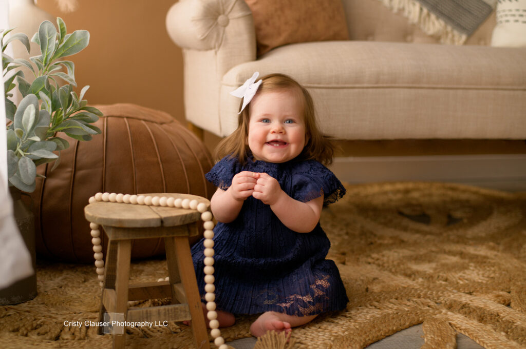 A smiling baby sitting on a woven rug, wearing a navy blue dress and a white bow in her hair. She is near a small wooden stool with a beaded necklace draped over it. The background includes a brown ottoman and a beige sofa with cushions.