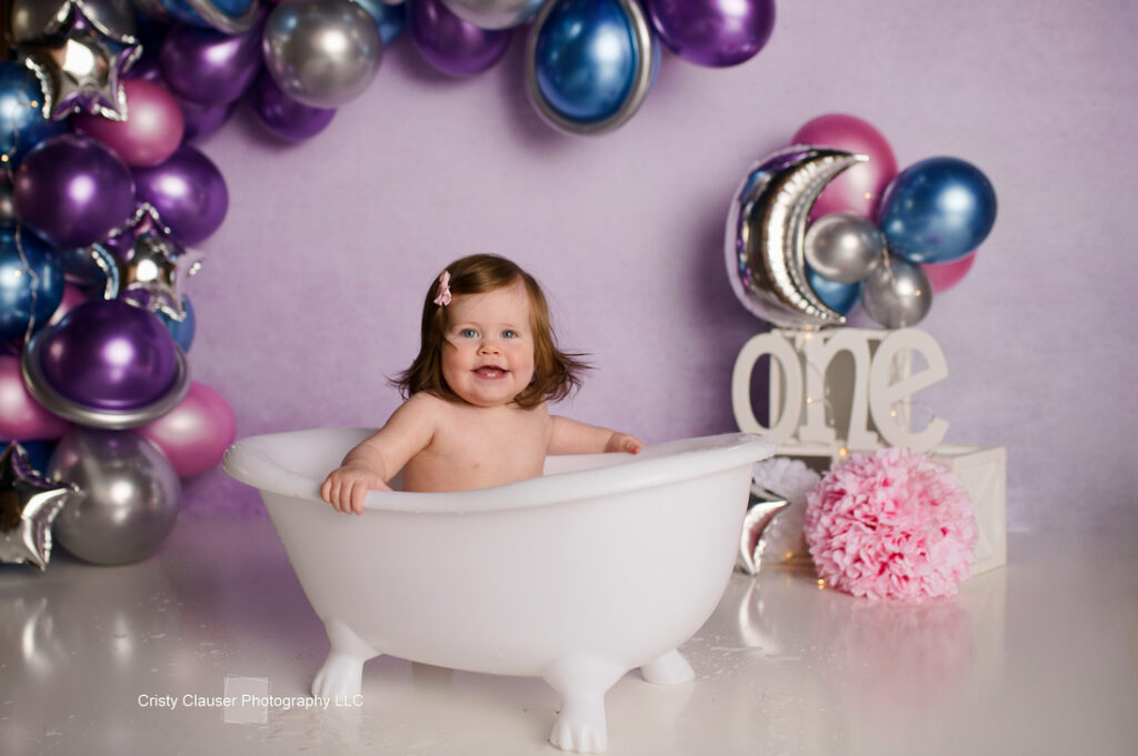 A smiling baby sitting in a white, mini clawfoot bathtub. The background features purple, blue, and silver balloons, with a large white number "one" to the side. A pink pom-pom decorates the floor.