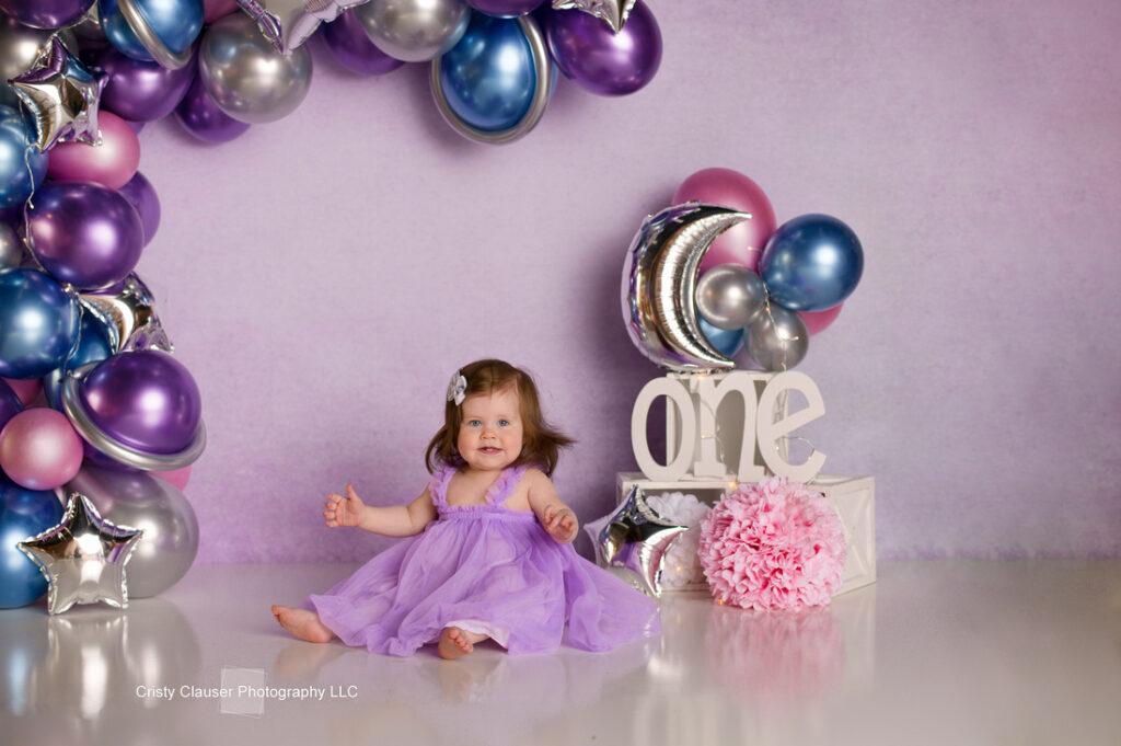A baby in a purple dress sits on the floor, surrounded by a festive balloon arch in blue, purple, and silver. A silver moon and the word "one" are next to her, with a pink fluffy decoration on the floor.