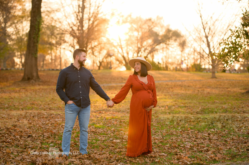 A couple stands in a sunlit park holding hands. The pregnant woman wears a long orange dress and a hat, smiling at her partner in a dark shirt and jeans. Autumn leaves cover the ground, and bare trees are in the background. Cristy Clauser Photography