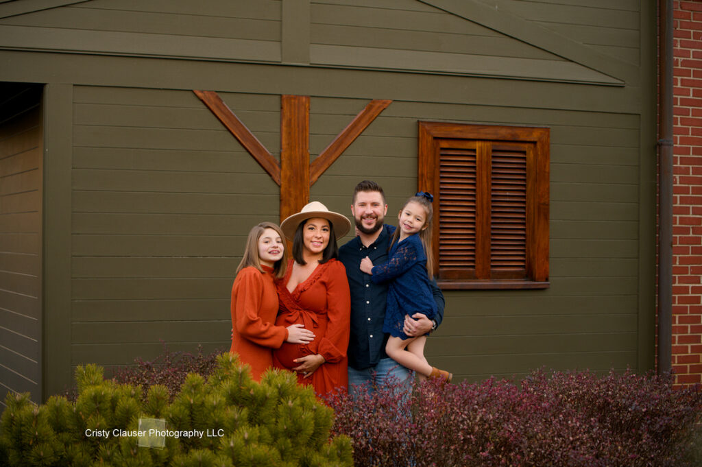 A family of four poses in front of a green building with wooden accents. The mother, pregnant and wearing an orange dress, stands next to the father in a navy shirt, with their two young daughters smiling beside them. Cristy Clauser Photography
