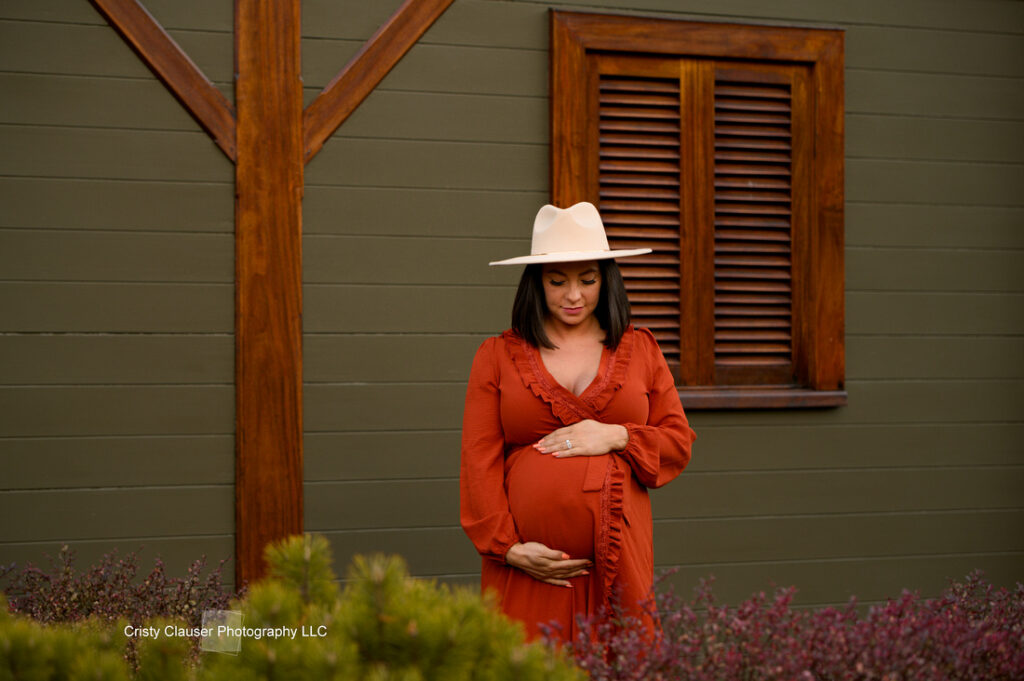 A woman in a rust-colored dress and wide-brim hat stands in front of a green building with wooden trim, holding her pregnant belly and looking down softly, surrounded by greenery. Cristy Clauser Photography