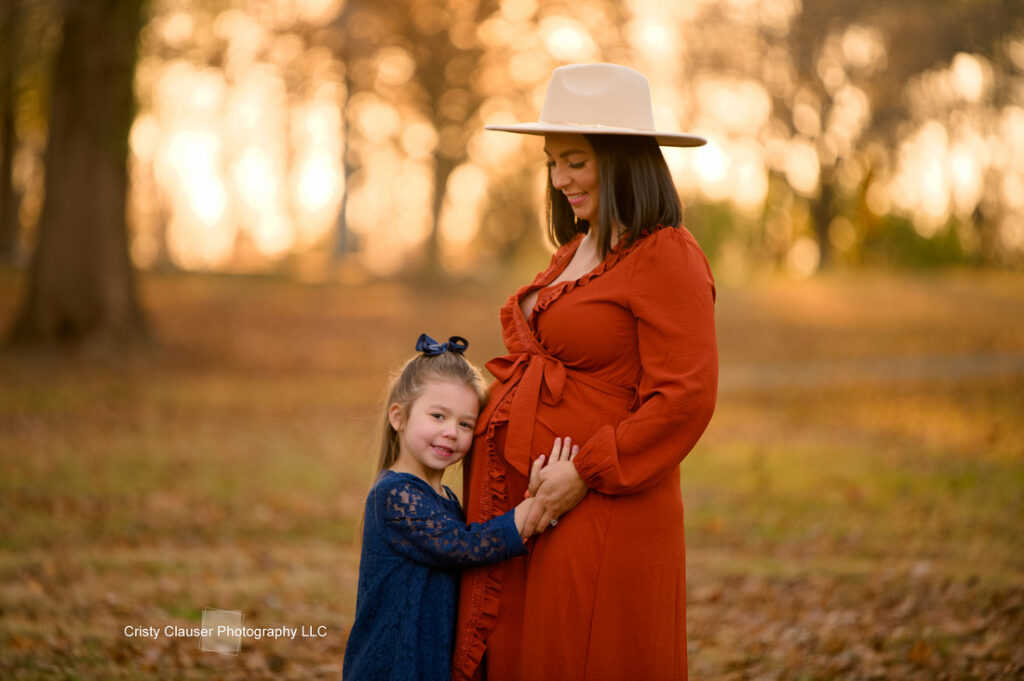 A pregnant woman in an orange dress and hat stands outdoors in an autumn setting, smiling as a young girl in a blue dress hugs her belly, both looking happy. Cristy Clauser Photography