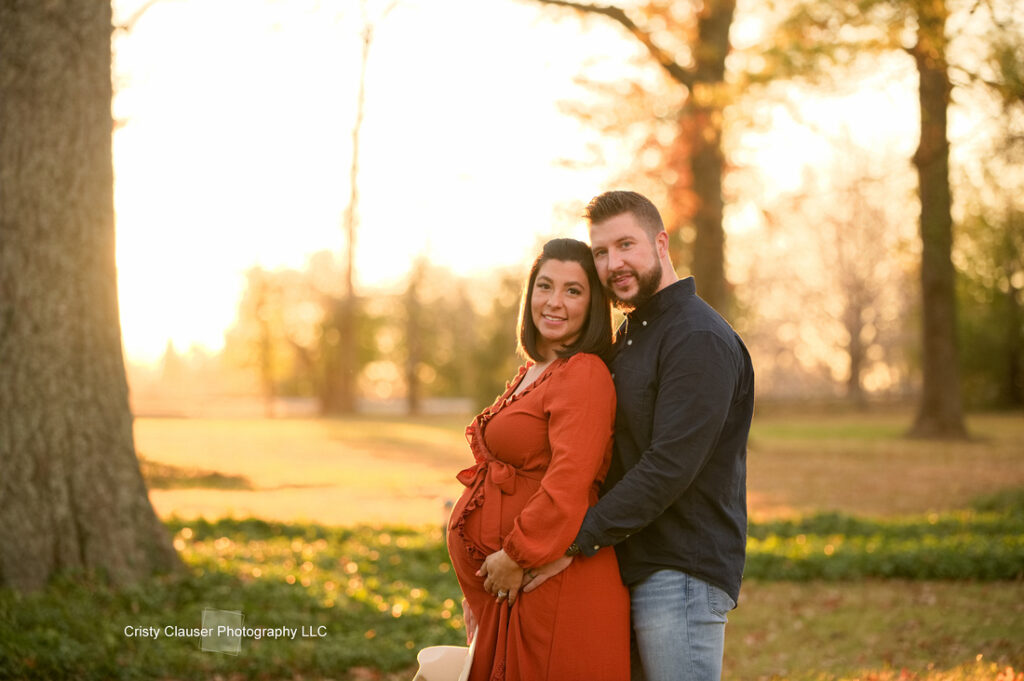 A couple poses outdoors in a sunlit park. The woman, wearing an orange dress and visibly pregnant, stands in front while the man hugs her from behind. Both smile at the camera. Autumn trees and golden light fill the background. Cristy Clauser Photography