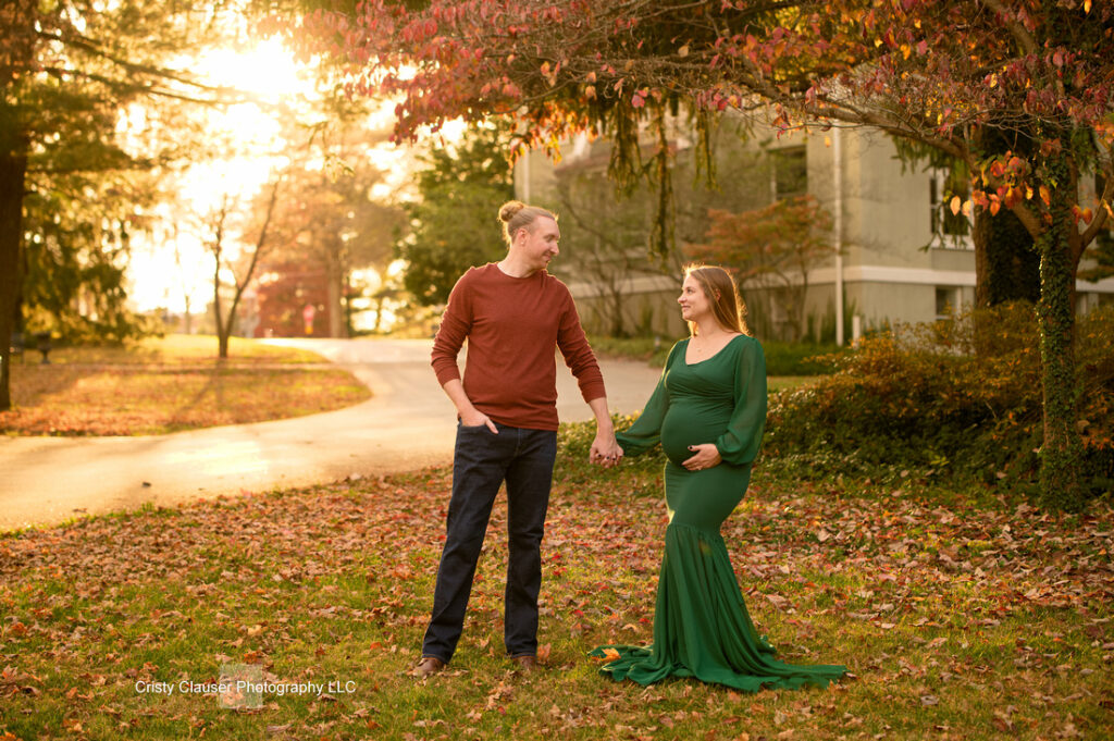 A pregnant woman in a long green dress holds hands with a man in a rust sweater as they stand on grass surrounded by autumn leaves, looking at each other, with golden sunlight in the background. Cristy Clauser Photography