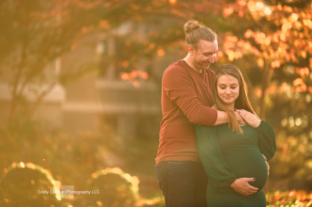 A man stands behind a pregnant woman in a green dress, gently embracing her as she holds her belly. They are outdoors with warm sunlight and autumn foliage in the background. Cristy Clauser Photography