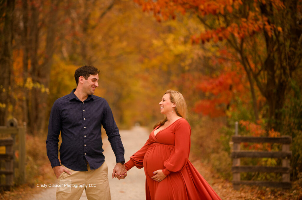 A pregnant woman in an orange dress holds hands with a man in a blue shirt as they smile at each other on a leaf-covered path lined with autumn trees. Cristy Clauser Photography