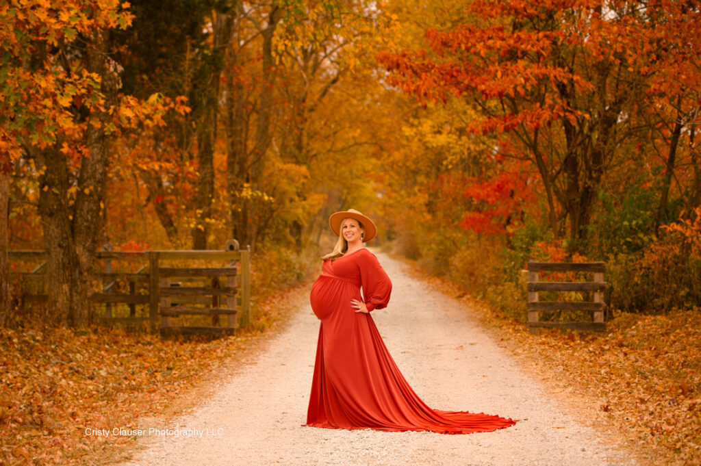 A pregnant woman in a flowing red dress and wide-brimmed hat stands on a tree-lined gravel path covered in autumn leaves, surrounded by vibrant orange foliage. Cristy Clauser Photography