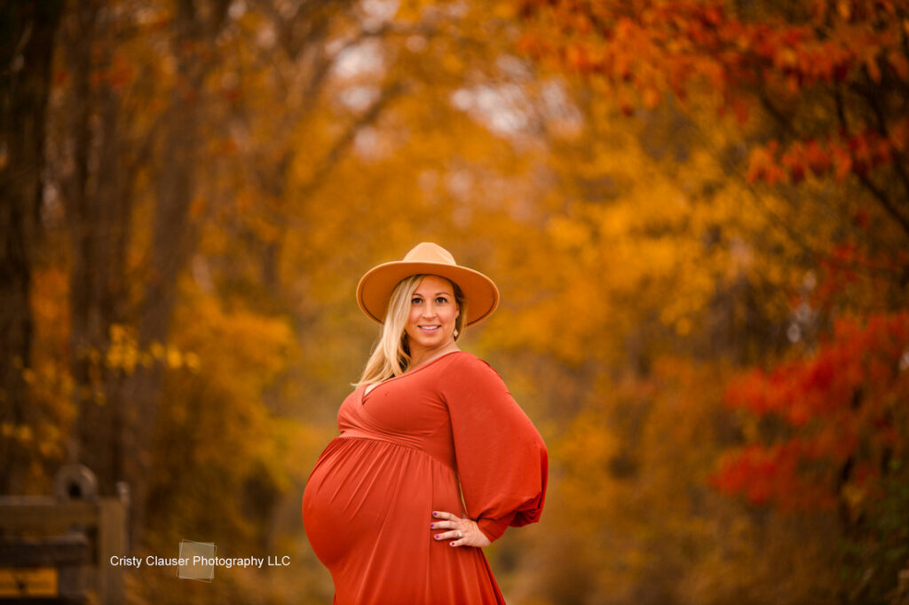 A pregnant woman in an orange dress and tan hat stands outdoors, smiling, surrounded by vibrant autumn trees with golden and red leaves. Cristy Clauser Photography