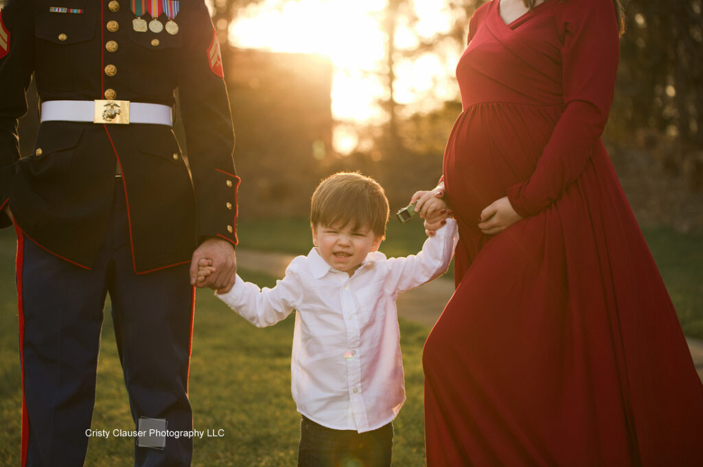A young boy stands between a man in a military uniform and a pregnant woman in a red dress, holding their hands outdoors at sunset. The boy squints and smiles toward the camera. Cristy Clauser Photography