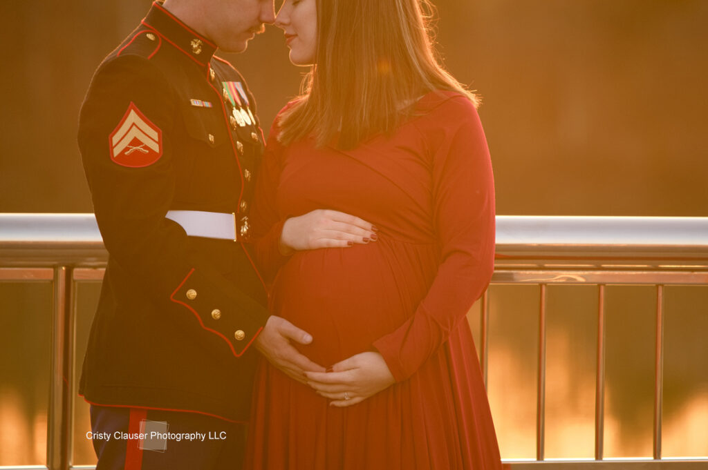 A man in a U.S. Marine Corps dress uniform and a pregnant woman in a red dress stand close, touching foreheads and holding her baby bump, bathed in warm, golden light by a railing. Cristy Clauser Photography