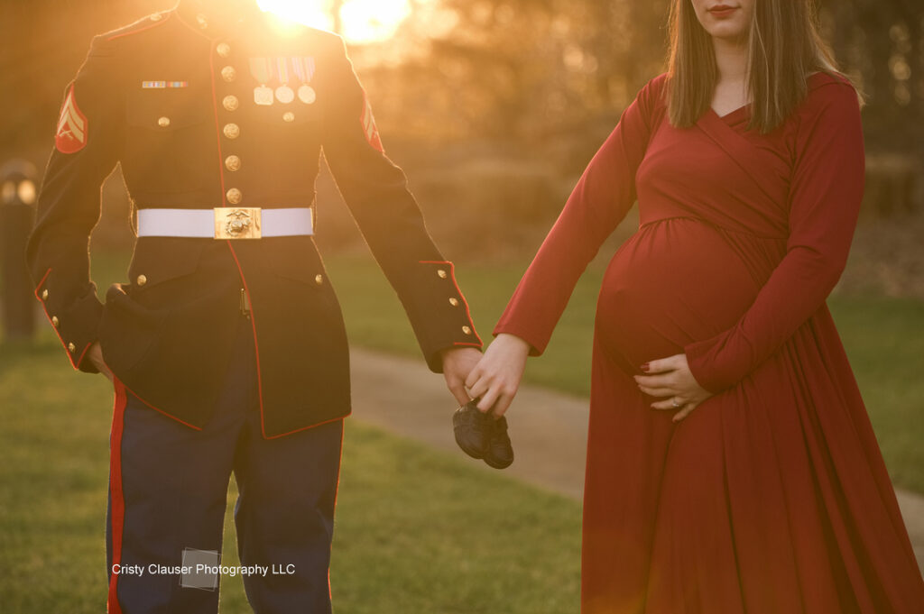 A Marine in uniform holds hands with a pregnant woman in a long red dress at sunset; she holds her belly and they hold tiny black baby shoes together, suggesting they are expecting a child. Cristy Clauser Photography