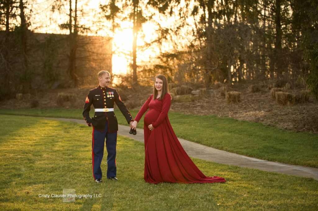 A man in a military dress uniform holds hands with a woman in a long red dress as they stand on grass at sunset, with trees and hay bales in the background. Cristy Clauser Photography
