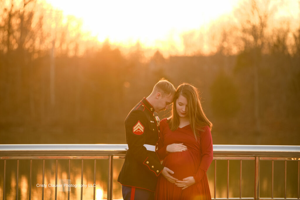 A U.S. Marine in dress uniform embraces a pregnant woman in a long red dress, resting his head gently on hers. They stand on a bridge at sunset, with soft golden light in the background. Cristy Clauser Photography