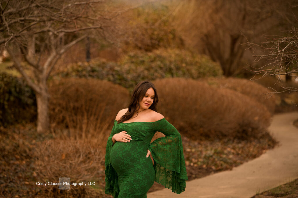 A pregnant woman in a long, off-the-shoulder green dress stands outdoors on a winding path, surrounded by bare trees and brown bushes, gently cradling her belly. Cristy Clauser Photography