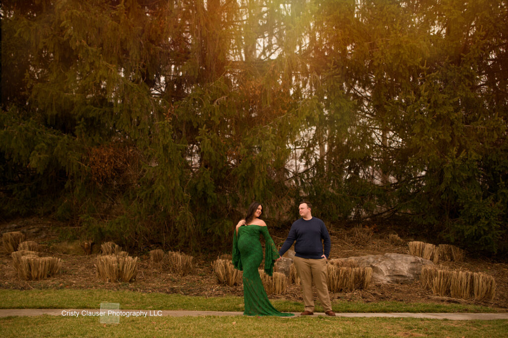 A woman in a long green dress holds hands with a man in a blue sweater as they stand on a sidewalk in front of tall evergreen trees. The scene is outdoors with sunlight filtering through the trees. Cristy Clauser Photography