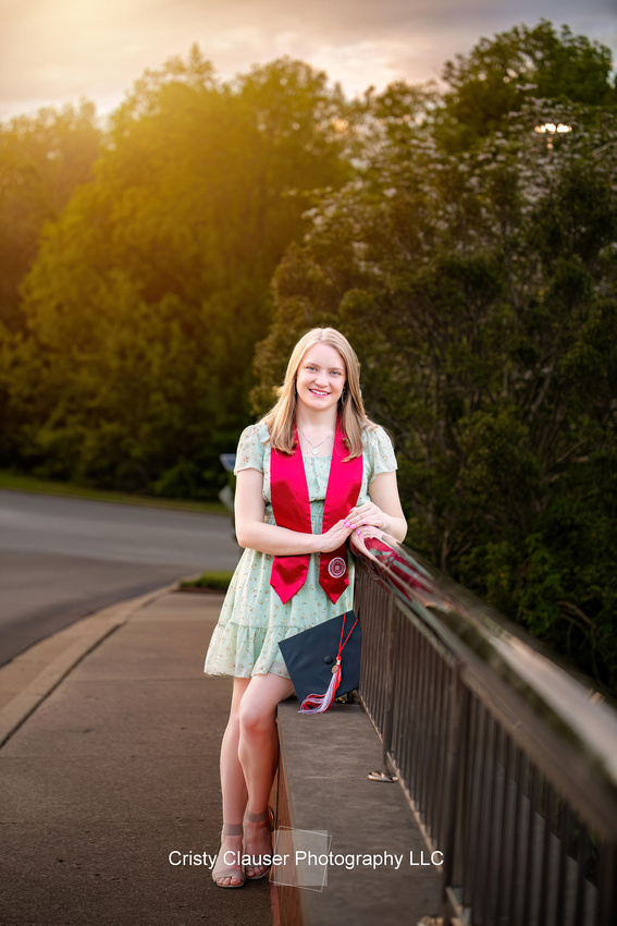 Young woman in a light floral dress and red stole stands on a bridge, leaning on the railing. She holds a graduation cap and smiles, with trees and a road in the background. Sunlight filters through the trees.