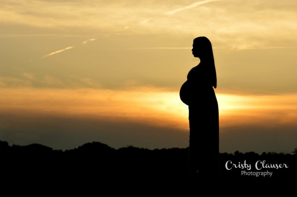 Silhouette of a pregnant woman standing outdoors at sunset, with a golden sky and dark landscape in the background. "Cristy Clauser Photography" is written in the lower right corner. Cristy Clauser Photography