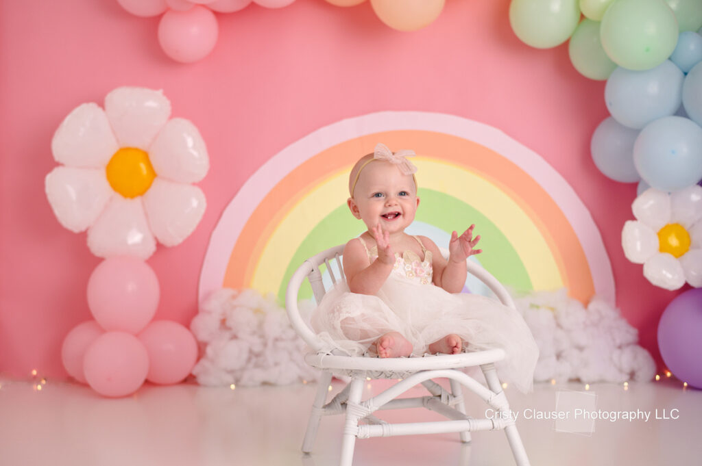 A smiling baby in a white dress sits on a small white chair. The background features a colorful rainbow mural with pink, blue, and white balloons and floral decorations. The setting is cheerful and bright.