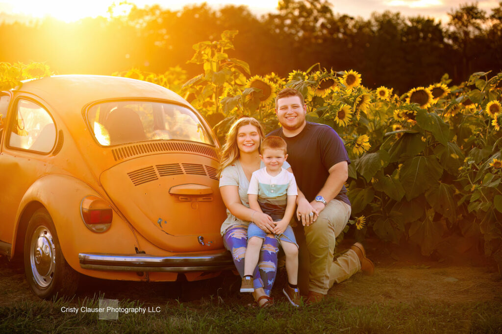 A smiling family of three sits beside an orange vintage car in a sunflower field at sunset, surrounded by bright yellow sunflowers and warm golden light. Cristy Clauser Photography