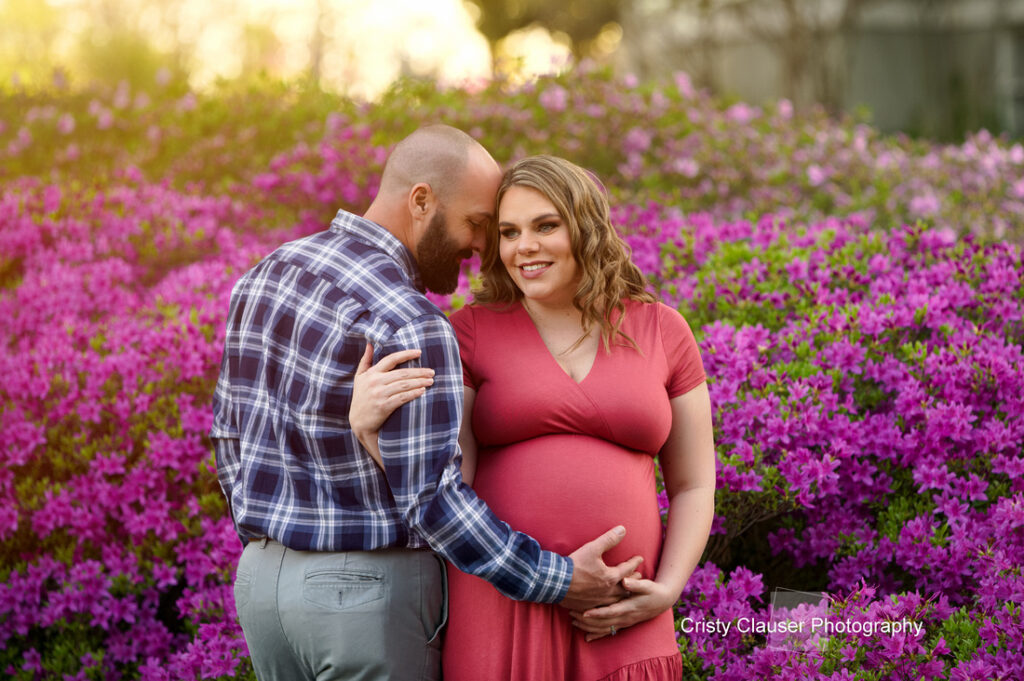 A smiling pregnant woman in a coral dress stands in front of vibrant pink flowers, while a man in a plaid shirt gently embraces her and touches her baby bump. They look happy and content outdoors. Cristy Clauser Photography