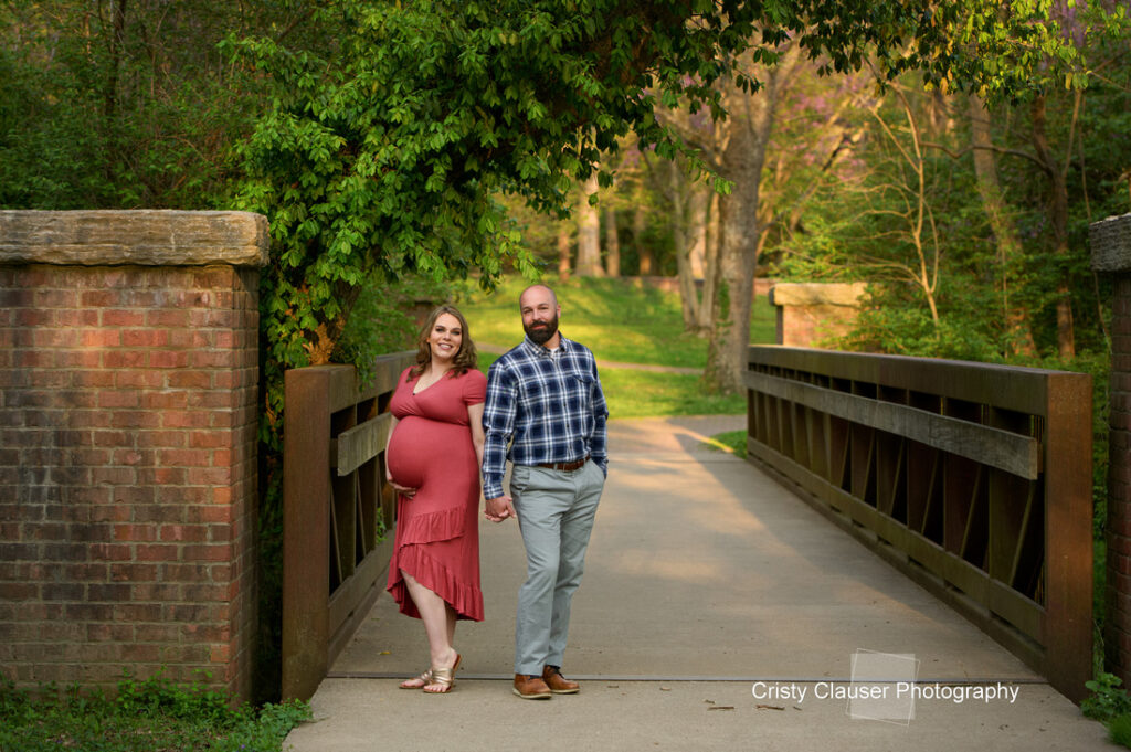 A pregnant woman in a coral dress and a man in a blue plaid shirt walk hand in hand on a wooden bridge surrounded by green trees in a sunlit park. Cristy Clauser Photography