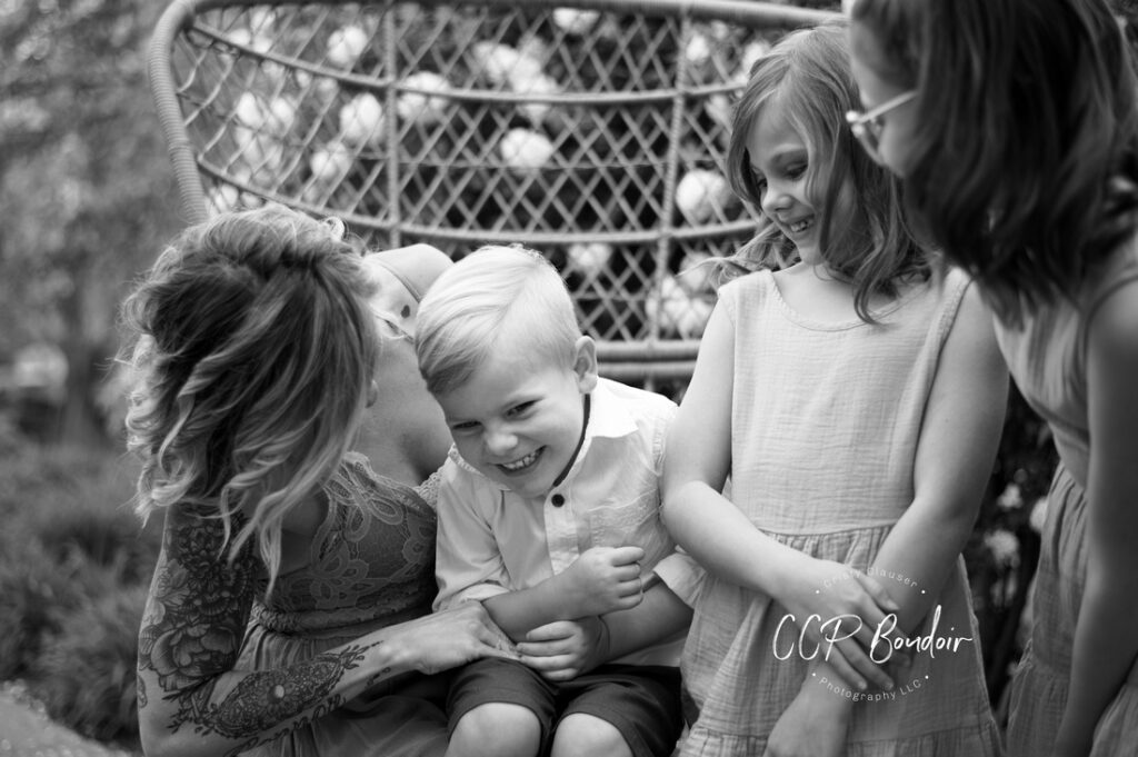 A woman playfully kisses a smiling young boy on the cheek while two girls stand beside them, all laughing together. They sit on a woven outdoor bench, sharing a joyful moment. Cristy Clauser Photography
