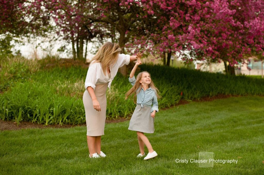 A woman and a young girl dance together on green grass, smiling under blooming pink trees. The woman twirls the girl, both wearing light-colored skirts and white shoes. The setting is vibrant and outdoor. Cristy Clauser Photography