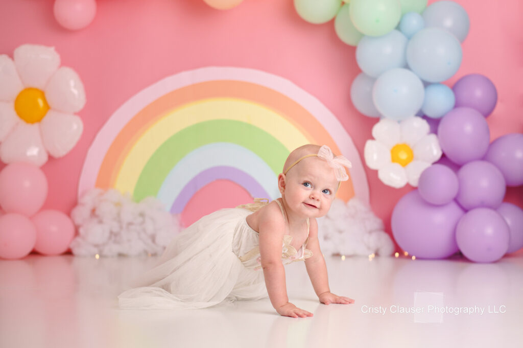 A smiling baby in a white dress and pink headband crawls on a white floor. Behind, there's a pastel rainbow, clouds, and colorful balloons in pink, purple, and blue against a pink background.