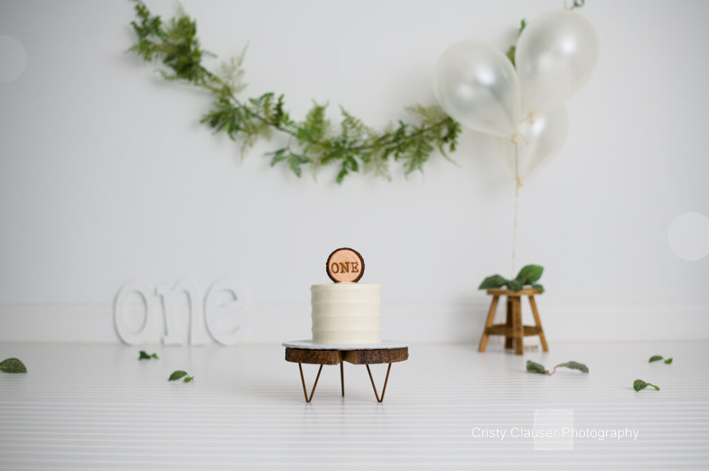 A small, white cake on a wooden stand is topped with a "ONE" sign. Green garland hangs in the background with white balloons tied to a small stool. The word "one" is displayed on the floor. The setting is minimal with a clean, white backdrop.