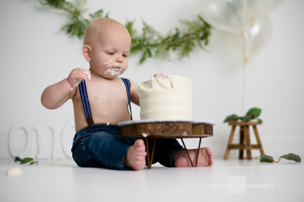 A baby with minimal hair sits on the floor, wearing blue jeans and suspenders. The baby is playfully exploring a white frosted cake on a small wooden stand. Green garland and balloons decorate the background.