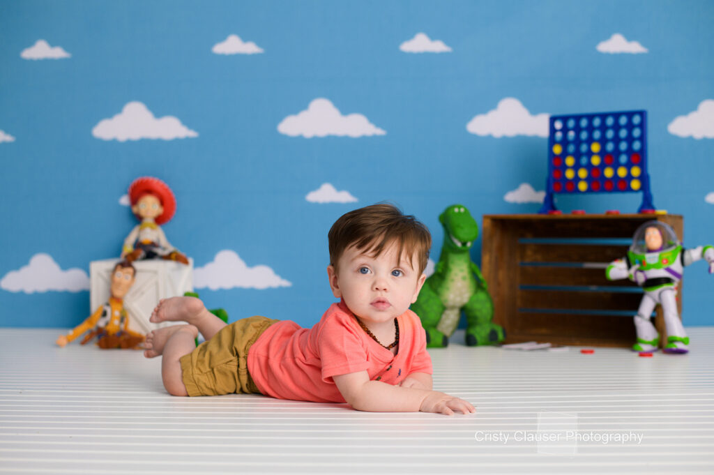 A baby in a coral shirt and tan shorts lies on a white floor with a Toy Story-themed backdrop featuring Woody, Jessie, Rex, and Buzz Lightyear toys, with clouds and a Connect 4 game in the background. Cristy Clauser Photography