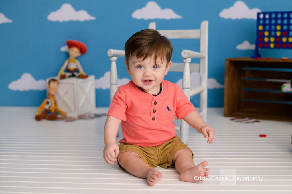 A smiling baby with brown hair sits on a white floor in front of a blue wall with white clouds. Toy Story characters Woody and Jessie are in the background, along with a Connect Four game. Cristy Clauser Photography