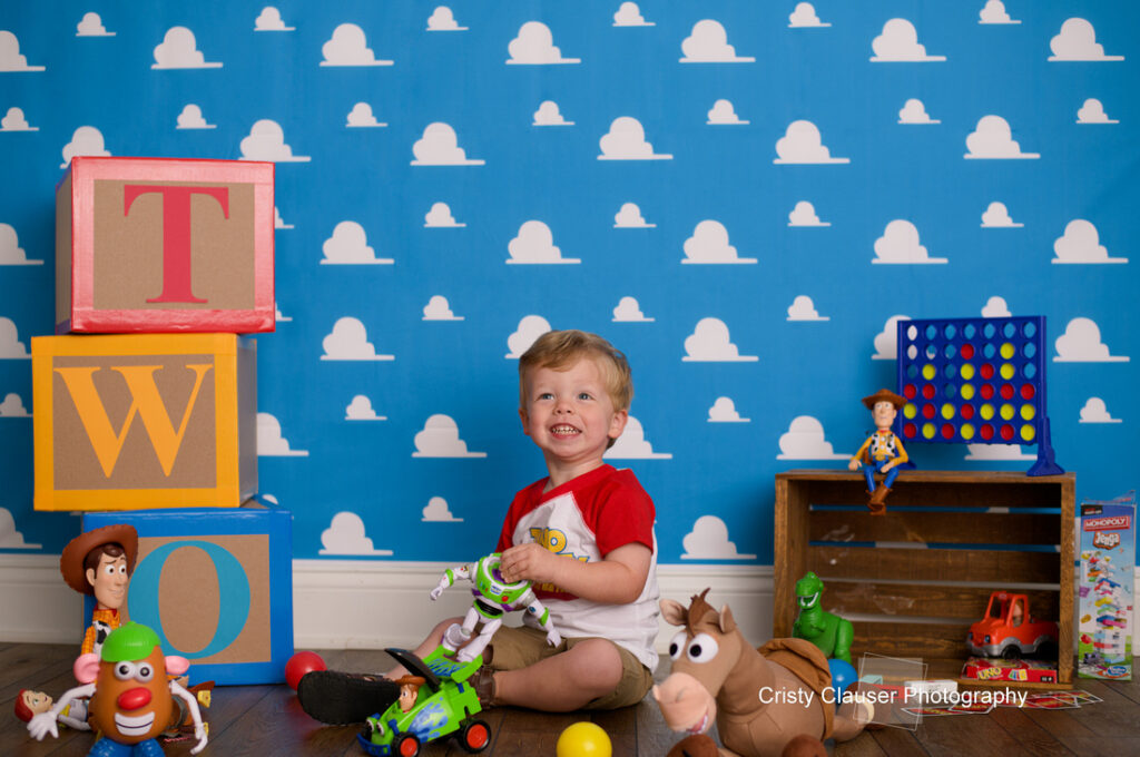 A smiling toddler sits on the floor with Toy Story toys and blocks spelling "TWO" against a blue wall with white clouds, celebrating a second birthday. Cristy Clauser Photography