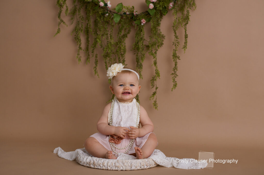 A baby wearing a light pink outfit and a white headband sits on a cushion, smiling. She is adorned with pearl necklaces. Behind her, green foliage and pink flowers are arranged decoratively. The background is a soft brown.