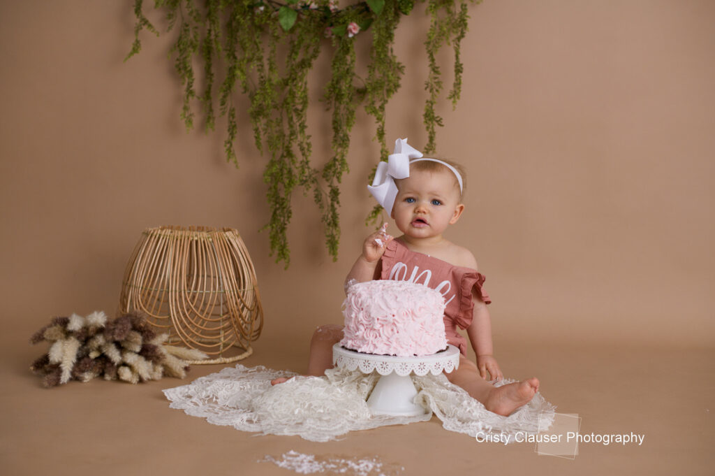 A baby in a pink outfit with a large white bow sits on the floor next to a pink frosted cake on a stand. Greenery and soft lighting decorate the background. A wicker lamp and dried flowers are on the side.