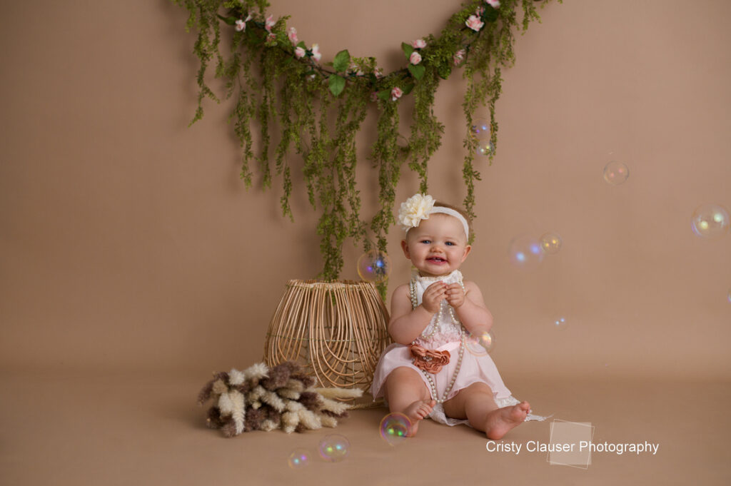 A smiling baby in a pink outfit with a floral headband sits on a neutral background, surrounded by bubbles. Behind her, there's a hanging arrangement of greenery and a basket with soft, fluffy materials. The photo is by Cristy Clauser Photography.
