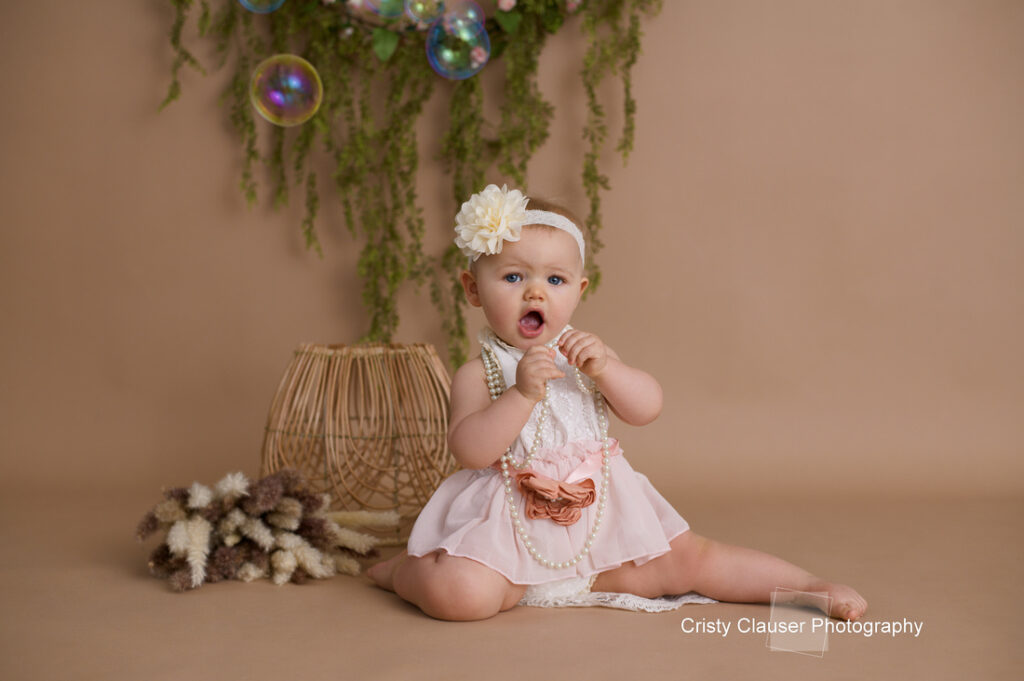 A baby wearing a light pink dress and headband with a white flower sits on a beige backdrop. Decorative plants with hanging greenery and bubbles surround her. She holds a string of beads and looks toward the camera.