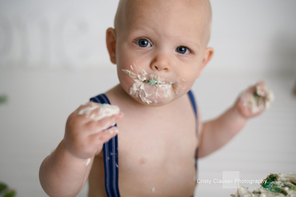 A baby with a shaved head and wide eyes is wearing blue suspenders. The baby has cake and frosting on their face and hands, appearing curious and playful. The background is blurred.