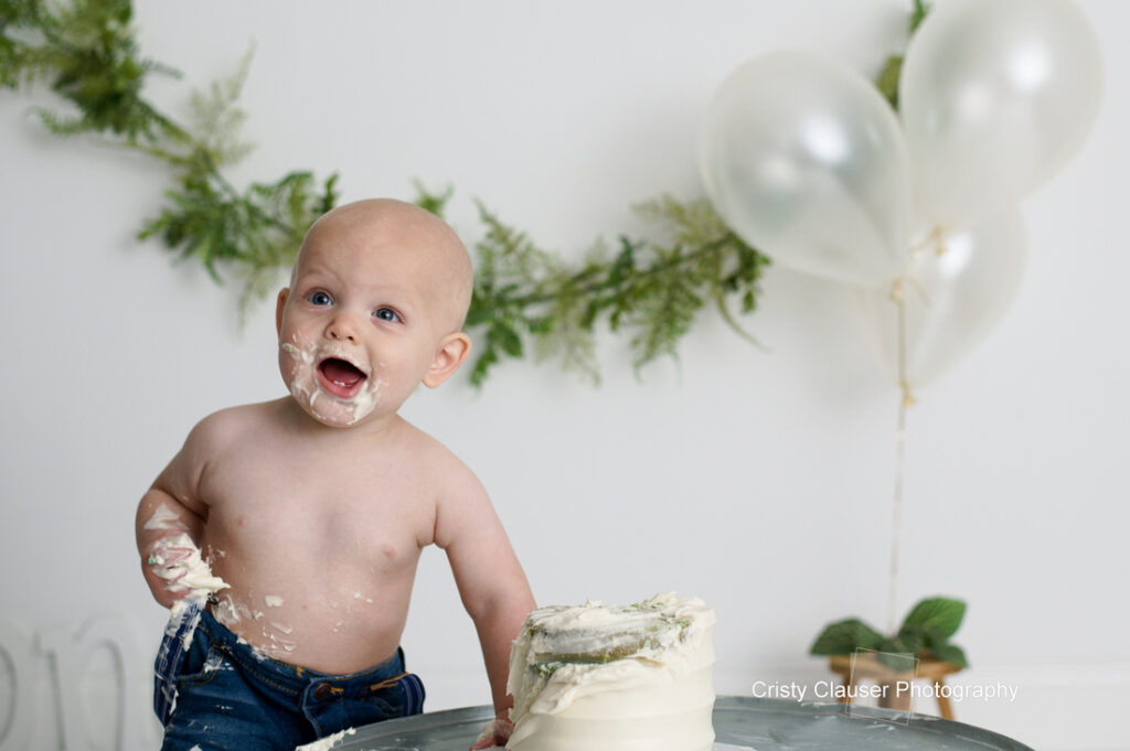 Baby with cake smeared on face and body, standing shirtless next to a white frosted cake on a table. Background has green leafy garland and white balloons, suggesting a celebration.